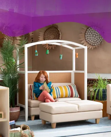 Girl sitting on a counch reading a book underneath the Sense of Place Cozy Classroom Arch with green potted plants around her