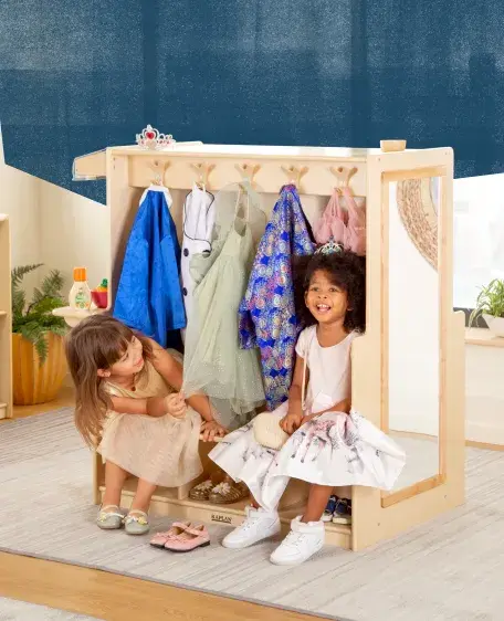 Two little girls sitting inside a dramtic play center with colorful costumes hanging on hooks above them